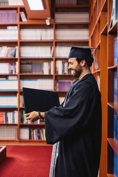 Emocionado graduado con una gorra y un vestido sosteniendo su licenciatura - Foto, imagen
