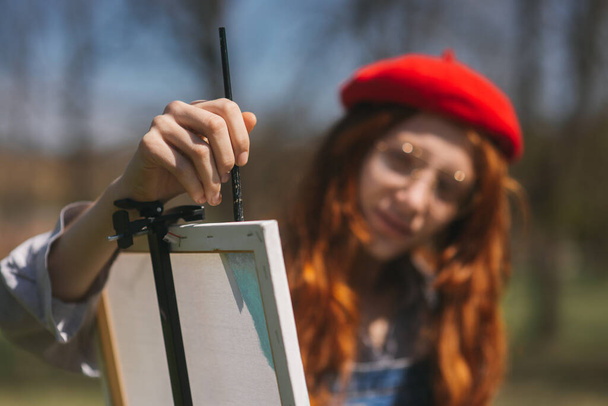 Κοντινό πλάνο του κορίτσι τζίντζερ με κόκκινο μπερέ ζωγραφική σε ένα λευκό καμβά στο πάρκο - Φωτογραφία, εικόνα