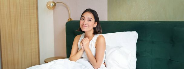 Портрет возбужденной, улыбающейся женщины, сидящей утром в своей постели, мечтающей, выглядящей обнадеживающей. - Фото, изображение