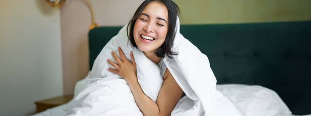 Женщина сидит на кровати, накрытая белым одеялом, улыбается, радуется счастливому уик-энду, смеется перед камерой. - Фото, изображение