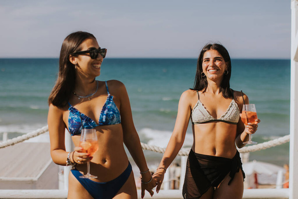 İki gülümseyen bikinili genç kadın kokteyl içerken sahilde tatilin tadını çıkarıyor. - Fotoğraf, Görsel
