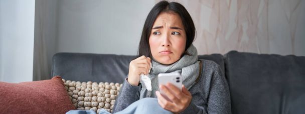 Азійська дівчина з грипом, сидить вдома, плаче і виглядає засмученою, тримає смартфон, почувається погано і похмуро, вдягнута в теплий одяг.. - Фото, зображення
