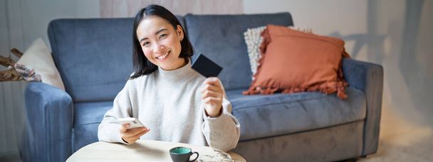 Mladá žena v obývacím pokoji, ukazuje svou kreditní kartu a pomocí smartphonu platit, objednat online nebo nakupovat na žádost. - Fotografie, Obrázek