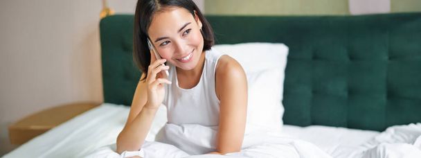 Smiling korean girl talks on mobile phone and lying in bed. Cute woman answers telephone call, holds smartphone, relaxing in her bed. - Photo, Image
