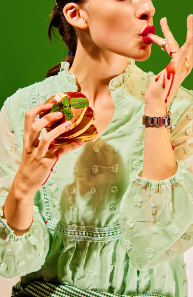 Śniadanie. Fotografia pop żywności. Kobieta trzymająca słodkie naleśniki, liżąca palce w dżemie. Vintage, lat 80-tych, styl lat 70-tych. Uzupełniające kolory. Pojęcie jedzenia, mody, stylu, smaku. Ogłoszenie - Zdjęcie, obraz