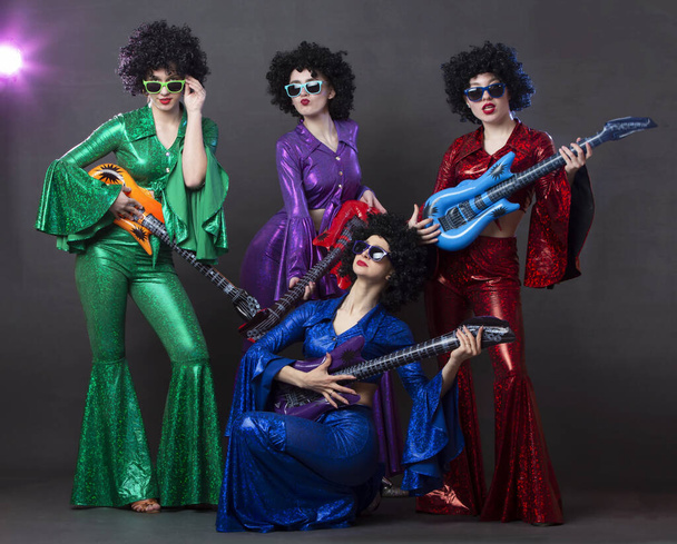 Egy popzenei csoport énekel egy nightclubban. Fiatal lányok öltözött retro hetvenes évek jelmezek és vicces afro parókák pózolnak együtt. Boldog nők szórakozás halloween diszkó - Fotó, kép