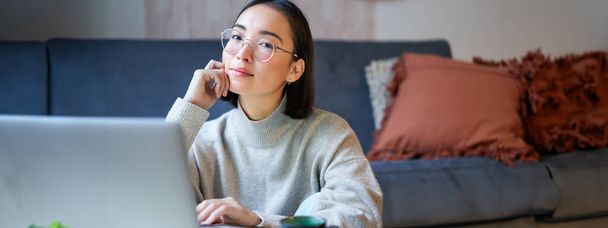 Zelfstandige jonge lachende vrouw, freelancer die thuis blijft, werkt op afstand van laptop, draagt een bril, zit in de woonkamer. - Foto, afbeelding