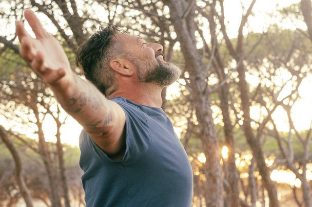 Side view of adult man with beard outstretching arms in outdoor leisure happy and free activity alone. One overjoyed male mature people excited in front of a sunset in the park. Concept of life joy - Photo, Image