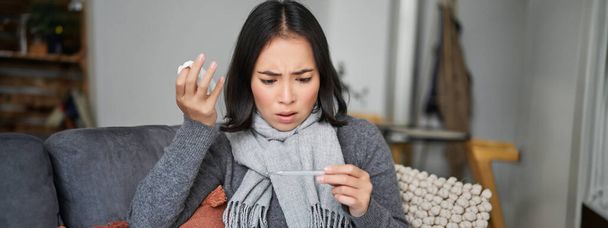 Woman looking concerned and shocked at thermometer, catching cold, feeling unwell with fever and covid symptoms, staying at home. - Photo, Image