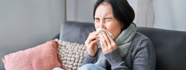 Close up of young korean woman staying at home with cold, sneezing in napking, has runny nose, concept of illness, health and influenza. - Photo, Image