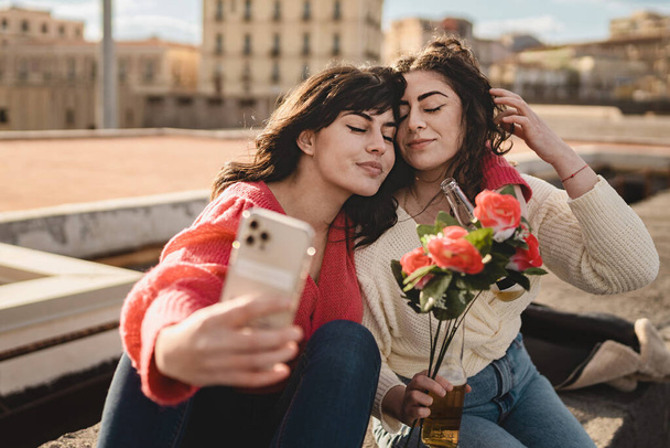 Una joven pareja de lesbianas de unos veinte años, tomando una selfie en un muelle, se acerca la cabeza con los ojos cerrados. Uno sostiene un ramo de rosas rojas. - Foto, Imagen
