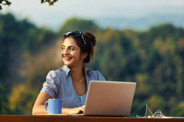 Femme avec les cheveux attachés, en utilisant des lunettes comme un bandeau, travaille à l'extérieur avec son ordinateur portable, au milieu de la verdure et des montagnes - Photo, image