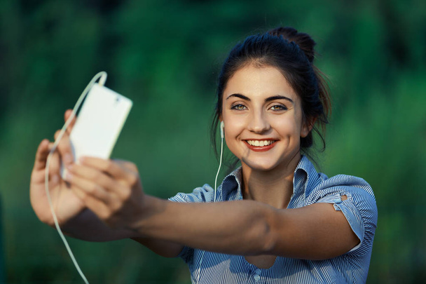 Die junge Frau im Freien nutzt das Smartphone für Unterhaltung, Selbstvermarktung, Influencer-Bemühungen, Content-Produktion und Selfies. Ihr Fokus: zugängliche und ansprechende Inhalte zu produzieren, sie zu präsentieren  - Foto, Bild