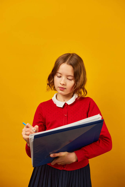 Adolescente réfléchie tenant un stylo et écrivant sur un carnet sur fond de studio jaune, concept de retour à l'école - Photo, image