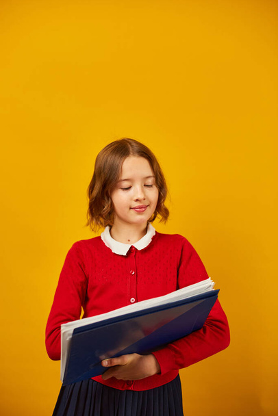 Задумчивая школьница-подросток держит ручку и пишет на блокноте на жёлтом фоне студии, возвращаясь к школьной концепции - Фото, изображение