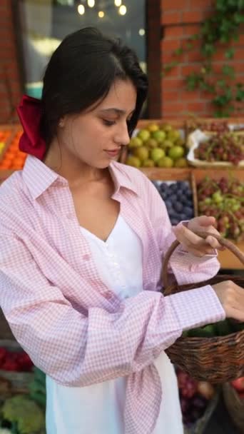 Kaunis nuori nainen sijoittaa vihannekset huolellisesti koriin Euroopan maanviljelijöiden markkinoilla. Laadukas 4k kuvamateriaalia - Materiaali, video