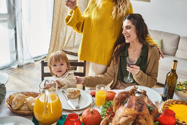 niedliche Kleinkind Mädchen Blick in die Kamera in der Nähe lgbt Eltern und Thanksgiving gebratenen Truthahn auf dem Tisch - Foto, Bild