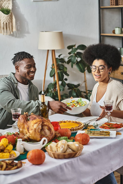 cheerful african american woman passing bowl with salad to relative during Thanksgiving dinner - Photo, Image