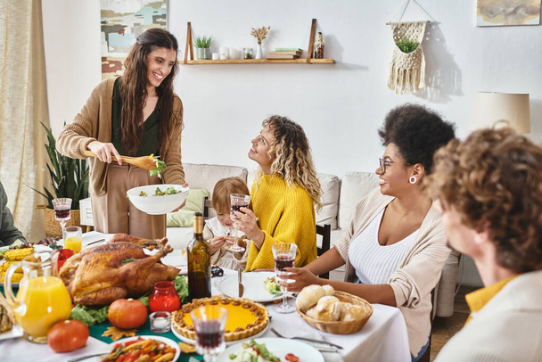 счастливая женщина, подающая салат для межрасовых друзей и семьи во время празднования Дня благодарения дома - Фото, изображение