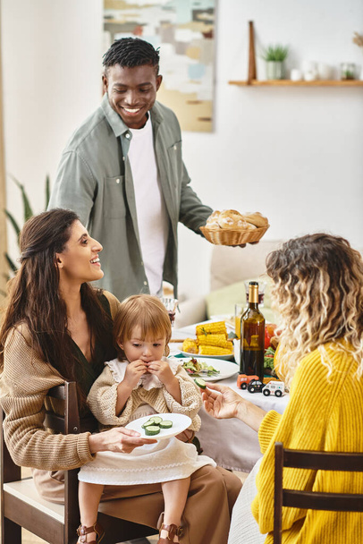Happy Thanksgiving, curly woman feeding toddler daughter with cucumber near multiethnic friends - Photo, Image