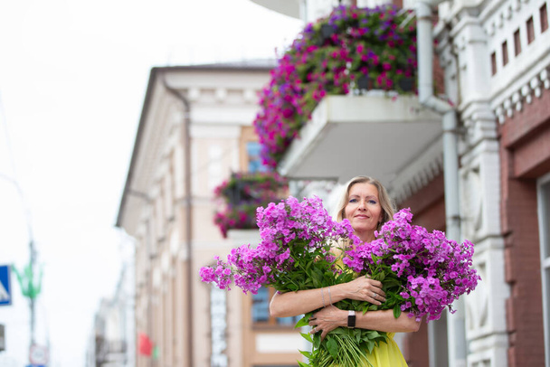 Une belle femme âgée avec un bouquet de fleurs se promène dans la ville. Modèle d'âge blond aux yeux bleus est heureux. Une femme de cinquante ans se promène en été, s'amuse. - Photo, image