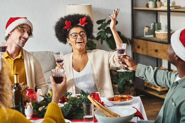 cheerful multicultural relatives in Santa hats raising glasses at festive table on Christmas - 写真・画像