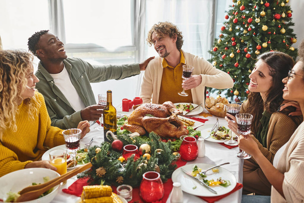Vielvölkige fröhliche Familie lacht und lächelt beim festlichen Mittagessen mit Weihnachtsbaum im Hintergrund - Foto, Bild