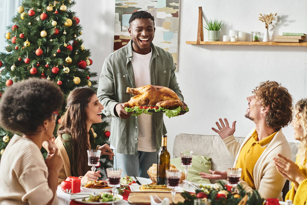 joyous african american surrounded by his multicultural family holding plate with turkey, Christmas - Foto, afbeelding