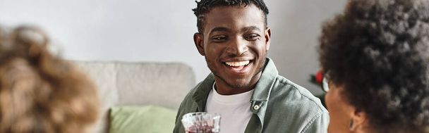alegre hombre afroamericano sentado en la mesa de Navidad sonriendo y levantando copa de vino, pancarta - Foto, imagen