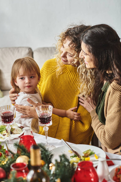 young lgbt couple in casual attire celebrating Christmas holding daughter in hands at festive table - Photo, Image