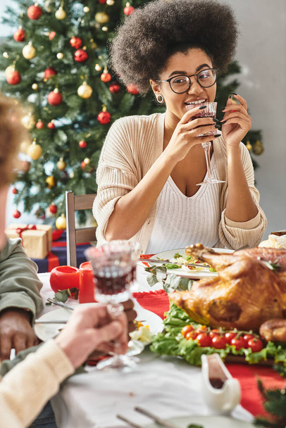 vrolijk Afrikaans amerikaanse vrouw praten met haar familieleden het drinken van wijn met kerstboom achtergrond - Foto, afbeelding