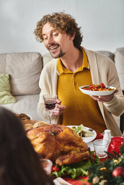 jonge vreugdevolle man zitten aan tafel met zijn gezin en genieten van feestelijk eten, Kerstmis - Foto, afbeelding