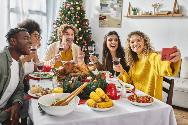 gran familia multirracial tomando selfie alegre sentado en la mesa festiva con árbol de Navidad en el telón de fondo - Foto, imagen