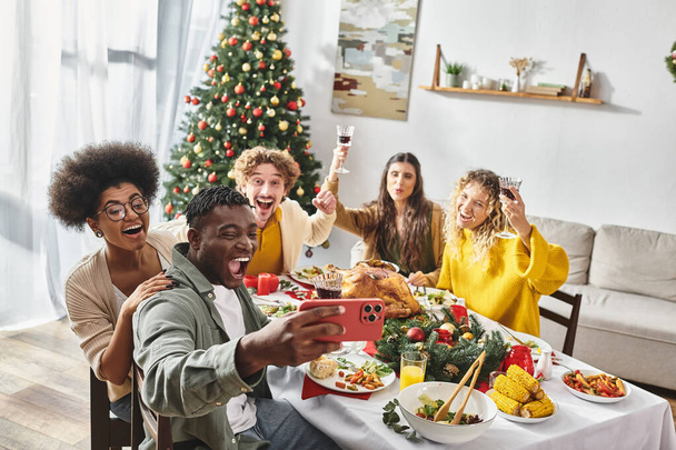 バックドロップのクリスマスツリーでお祝いのテーブルでセルフィーを取る楽しい多民族家族のメンバー - 写真・画像