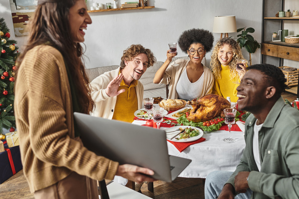 многонациональная счастливая семья, сидящая за праздничным обедом, радуясь вину и улыбаясь в камеру ноутбука - Фото, изображение