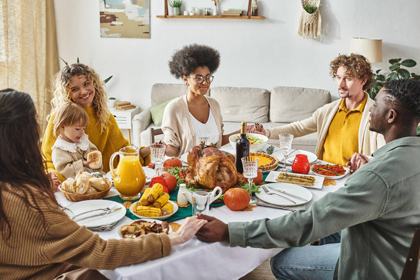 joyeux membres de la famille multiculturelle assis et tenant la main priant à la table festive, Thanksgiving - Photo, image
