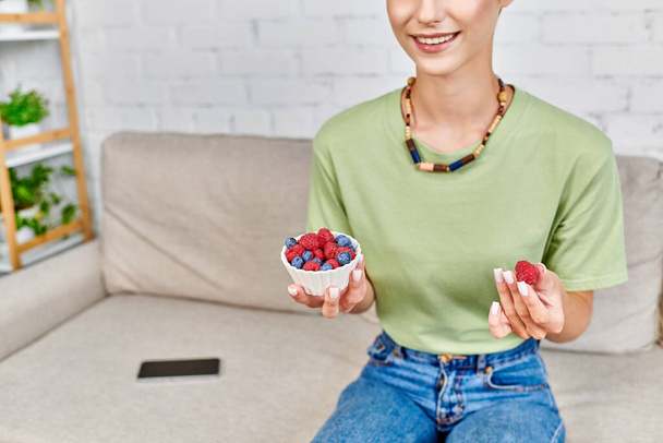 cropped view of vegetarian woman with bowl of fresh raspberries and blueberries in living room - Foto, afbeelding