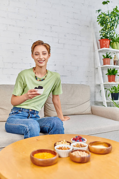cheerful woman with ripe blackberries near assortment of plant-based food on table in living room - Zdjęcie, obraz