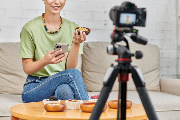 mujer recortada con teléfono inteligente y tazón de anacardos cerca de comida vegetariana durante el video blog en casa - Foto, Imagen