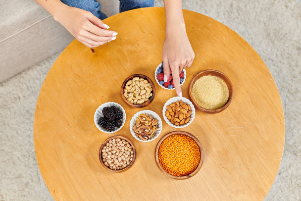 mujer recortada apuntando a la mesa con diversas bayas y frutos secos con legumbres, nutrición a base de plantas - Foto, Imagen