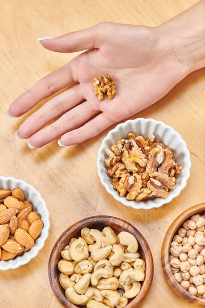 top view of female hand with walnut near almonds and cashews with chickpeas in bowls, vegetarianism - Foto, afbeelding