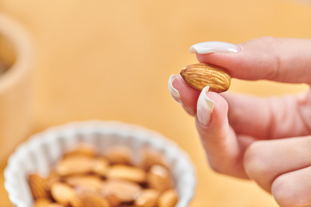 close up view of female hand with almond above bowl with vegetarian food on blurred background - Photo, Image
