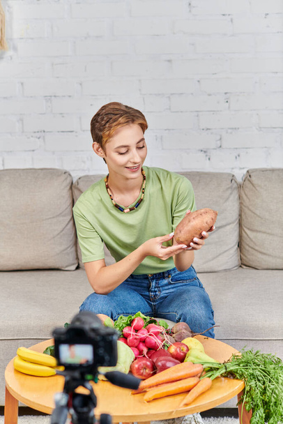 smiley woman with sweet potato near fresh vegetables and fruits in front of blurred digital camera - Photo, Image