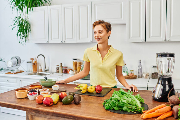 χαρούμενη γυναίκα κοντά στο τραπέζι με διάφορα συστατικά φυτικής προέλευσης και ηλεκτρικό μπλέντερ στη σύγχρονη κουζίνα - Φωτογραφία, εικόνα