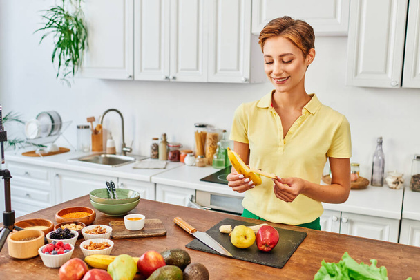 smiley woman peeling ripe banana near fruits and vegetables on table in kitchen, vegetarian concept - Foto, imagen