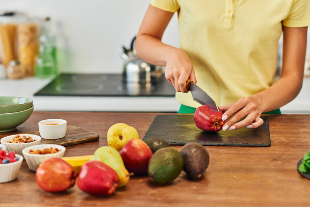 обрезанная женщина режет яблоко рядом со свежими фруктами и различными ингредиентами растительного происхождения, растительная диета - Фото, изображение