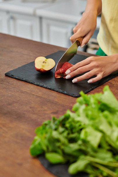 cropped view of woman cutting apple near fresh lettuce on worktop in kitchen, plant-focused diet - Фото, изображение