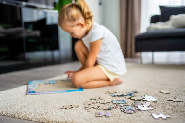 Little blonde girl sits at home on the carpet and collects puzzles. High quality photo - Photo, Image