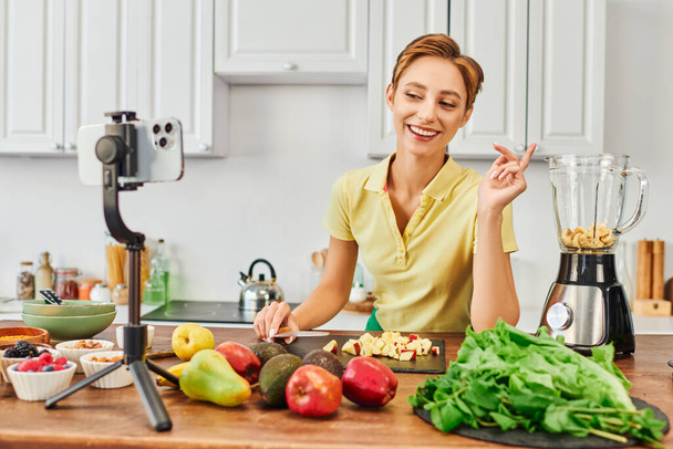 cheerful vegetarian woman looking at smartphone on tripod near fruits and electric blender, vlog - Photo, image