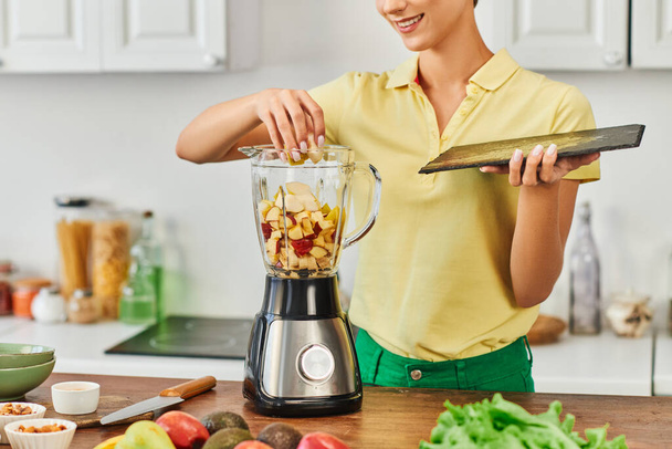 vista recortada de mujer alegre mezclando frutas picadas en licuadora eléctrica, deliciosa receta vegetariana - Foto, imagen